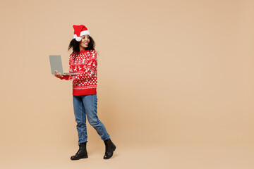 Full size body length smiling fun young african american Santa woman in Christmas hat sweater go move hold use work on laptop pc computer look back isolated on pastel beige background studio portrait.