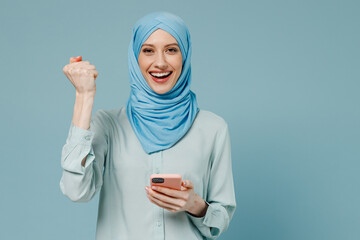 Young amazed happy arabian asian muslim woman in abaya hijab hold in hand use mobile cell phone do winner gesture isolated on plain blue background. People uae middle eastern islam religious concept