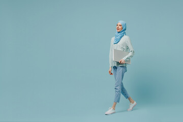 Full body side view young arabian asian muslim woman in abaya hijab hold closed laptop pc computer walking isolated on plain blue background studio. People uae middle eastern islam religious concept