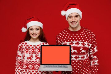 Young smiling couple friends two man woman in sweater hat hold use work on laptop pc computer blank screen workspace area isolated on plain red background studio portrait. Happy New Year 2022 concept.