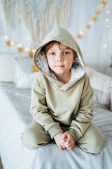 A boy in a light tracksuit sits on the bed. Behind him hangs a garland with stars.