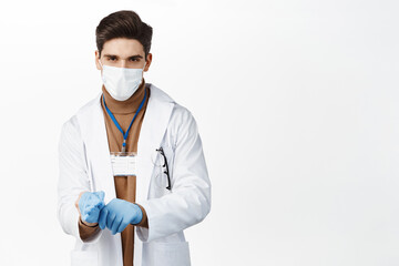 Covid 19. Doctor in medical face mask, put on gloves for patient examination, wearing clinic uniform, standing over white background