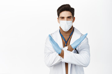 Young doctor in medical face mask from covid 19, rubber gloves, making cross stop gesture, prohibit action, standing over white background