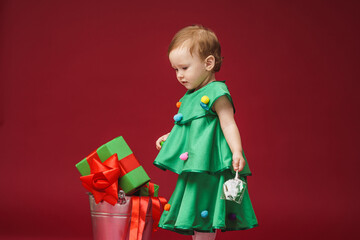 little girl in a Christmas tree costume and with a festively decorated headband is standing in a bucket on a red background. A child holds Christmas toys in his hands