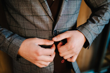 Man fastens the buttons jacket. The groom in a suit, shirt is standing on room background. Close up.
