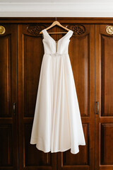 Fashionable beautiful classic lace silk wedding dress hanging on hanger in hotel wooden room. morning preparation wedding concept. vintage wedding gown.