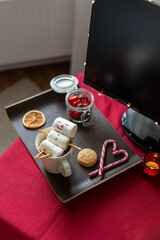 christmas, winter holidays and leisure concept - close up of marshmallow snowman taking bath in cup of coffee, sweet treats and tv monitor on table with red cloth at home