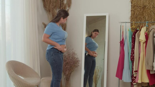 Young adult woman checking fat on her abdomen, standing in front of mirror at home. Woman expressing dissatisfaction of her shape, squeezing belly folds and feeling disappointment with body indoors