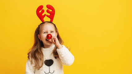 Happy little girl in fluffy white sweater with reindeer horns and a Christmas ball instead nose on yellow background. child is happy, having fun and preparing for Christmas. Advertising. Copy space.
