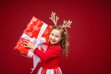 Happy little girl 4 years old in reindeer antlers on a bright red background. child is happy and holds gift in his hands and stretches it forward. Give gifts. New year sales, advertising, discounts.