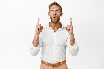 Portrait of handsome man pointing and looking up with wow face, checking out big sale, standing...