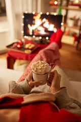 christmas, winter holidays and leisure concept - close up of young woman watching tv with fireplace on screen and holding mug of marshmallow and whipped cream resting feet on table at cozy home