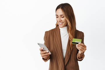 Stylish corporate woman using credit card and mobile phone to order online, paying with smartphone and smiling, standing over white background