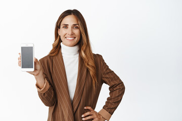 Confident saleswoman, middle aged woman showing smartphone empty screen, app interface, standing over white background