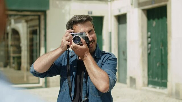 Front view of happy man taking picture of his lover in street. Slider shot of bearded Caucasian male person holding camera, smiling, taking photo during trip on summer day. LGBT, photoshoot concept