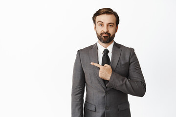 Unamused bearded male entrepreneur, wearing suit, pointing left and looking with disbelief, standing against white background