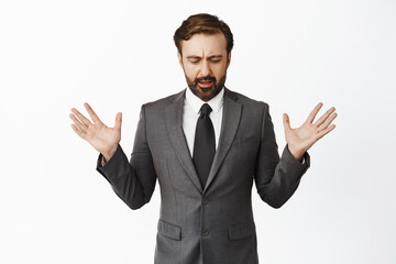 Confused businessman in suit raising empty hands up, looking down shocked, drop something below, standing over white background