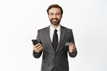 Business and technology concept. Handsome corporate man smiling pleased, holding credit card and smartphone, white background