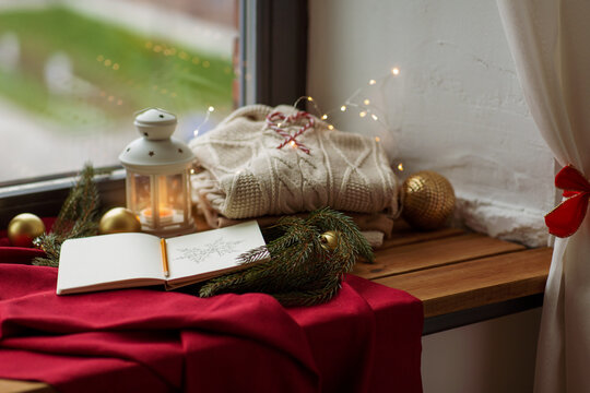 christmas, holidays and objects concept - close up of sketchbook with pencil drawing of snowflake, warm wool braided sweater, lantern and fir branch on red tablecloth on window sill at home