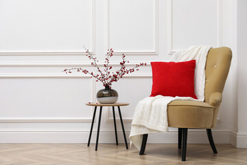 Hawthorn branches with red berries in vase on table near armchair indoors