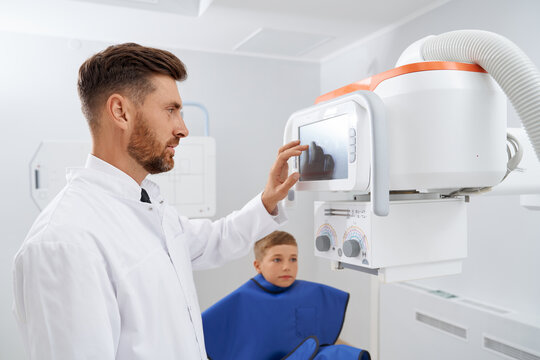 Side view of brunette doctor with beard touching screen of usg modern tool, starting procedure of diagnostic. Professional radiologist doing ultrasound diagnostic for little boy wearing blue robe. 
