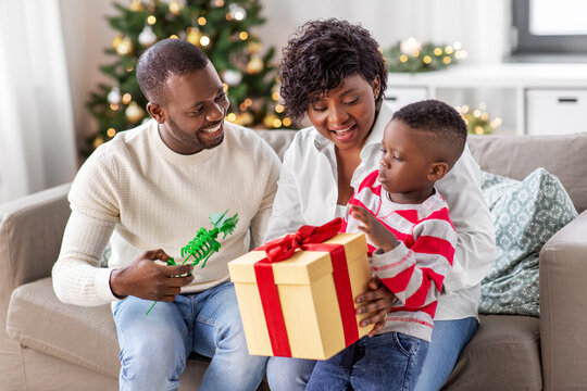 family, winter holidays and people concept - happy african american mother, father and baby son with gift box toy and toy at home on christmas