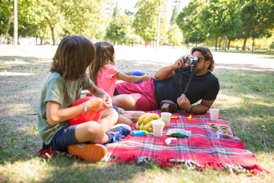 Loving father with disability on picnic with children. Man with mechanical leg in shorts lying on blanket with little boy and girl, inflating balloons, taking photos. Disability, family, love concept