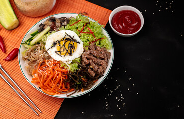 bibimbap or pibimpap, one of the most popular dishes of Korean cuisine. Boiled rice with various salads and egg. Serving with broth and sauce on a bamboo orange napkin, close-up on a black background