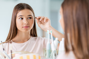 beauty, make up and cosmetics concept - teenage girl with tweezers tweezing her eyebrow and looking to mirror at home bathroom