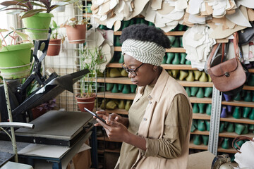 African dressmaker talking online with her customers using mobile phone while standing in the workshop
