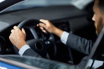 Side view of bearded businessman in stylish suit holding hands on steering wheel while sitting inside modern car. Male client buying new auto at salon.