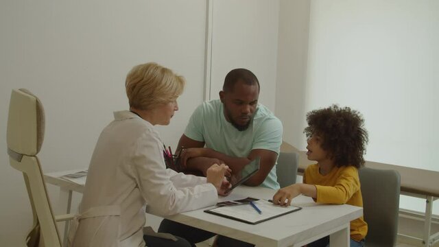 Pretty middle aged female pediatrician writing digital prescription on tablet pc, explaining treatment plan of sick cute elementary age african american boy to worried father in medical office.