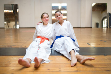 Happy young female karatekas sitting and hugging together. Cheerful Caucasian girls resting after karate class in studio. Sports, martial arts, healthy lifestyle concept © KAMPUS
