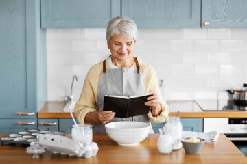 culinary, baking and people concept - happy smiling woman with cook book cooking food on kitchen at home