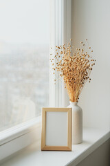 Photo frame mockup and vase with dried flax flowers on windowsill. Scandi style living room interior decorations.