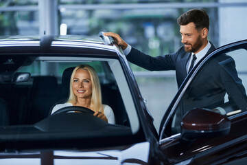 Fototapeta na wymiar Beautiful young woman with blond hair choosing new car at salon with help of professional salesman. Bearded man in suit working with potential female client at showroom. 