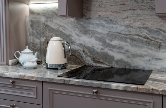 beautiful closeup of custom designed kitchen, with marble looking quartz countertop and backsplash. cream electric kettle with porcelain tea accessories on the marble countertop next to ceramic hob