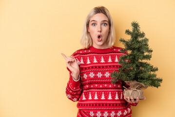 Young caucasian woman holding a little christmas tree isolated on yellow background pointing to the side