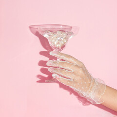 Creative layout with hand in lace glove holding martini cocktail glass with diamonds and pearls on pastel pink background. 80s or 90s retro aesthetic fashion concept. Romantic Valentines day idea.