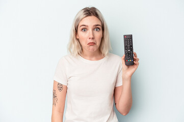 Young caucasian woman holding a tv controller isolated on blue background shrugs shoulders and open...