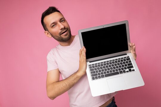 Handsome brunet man holding laptop computer with blank black monitor for mockup looking at camera in pink t-shirt isolated on pink background with free space for text