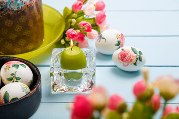 Easter composition with decorated eggs, burning candle, Easter bread or cake with glaze and multicolored sugar sprinkles. Unfocused flowers on foreground, copy space.