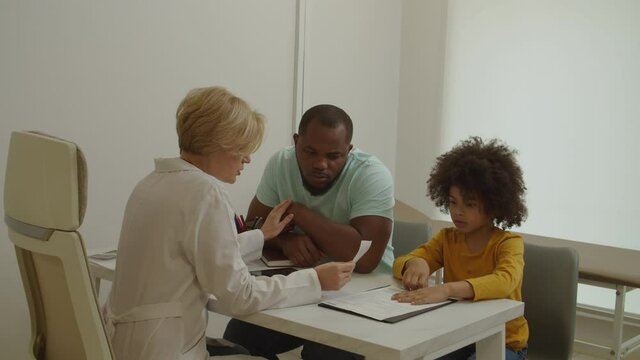 Empathetic professional woman doctor telling bad news to upset black father about illness of little son, showing medical test results, expressing support and solace at medical office.
