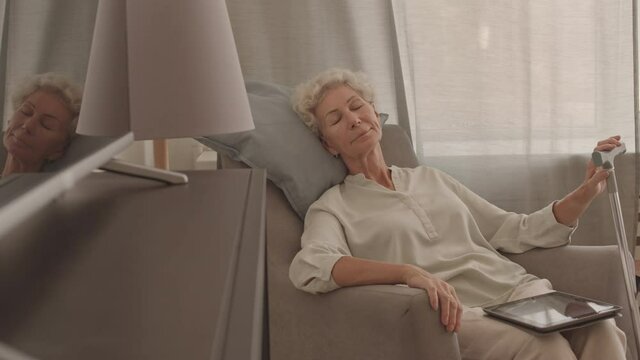 Stab medium shot of relaxed senior woman napping at home, sitting in armchair by window, holding digital tablet on her laps and walking stick in hand