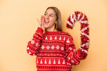 Young caucasian woman holding big christmas stick isolated on yellow background shouting and holding palm near opened mouth.