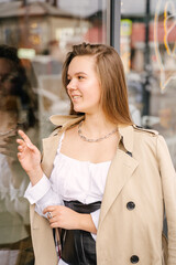 A young woman in a beige raincoat stands near a shop window