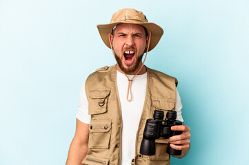 Young caucasian man looking at animals through binoculars isolated on blue background screaming very angry and aggressive.