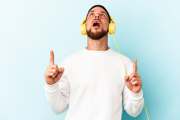 Young caucasian man listening to music isolated on blue background pointing upside with opened mouth.