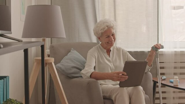 Stab medium shot of cheerful modern old lady video chatting on digital tablet sitting in soft armchair at cozy apartment