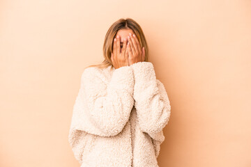 Young caucasian woman isolated on beige background blink at the camera through fingers, embarrassed covering face.